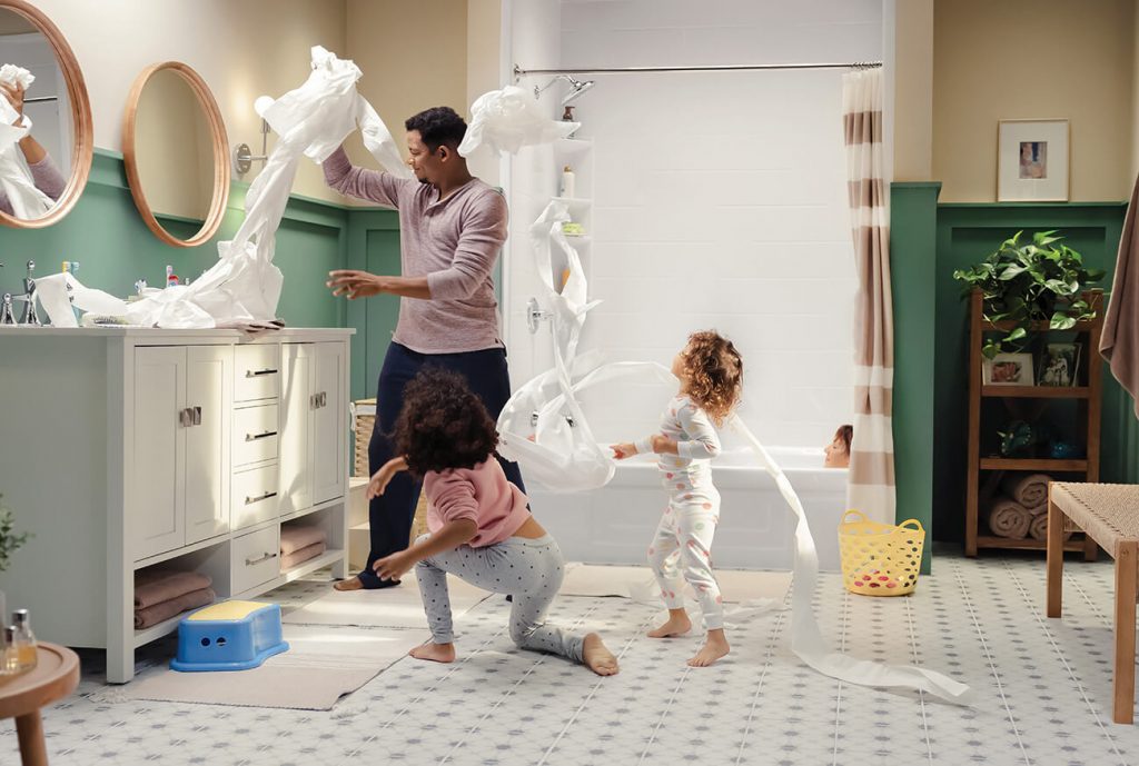 A mom takes a relaxing bath in the tub, while dad and kids are playing in the bathroom.