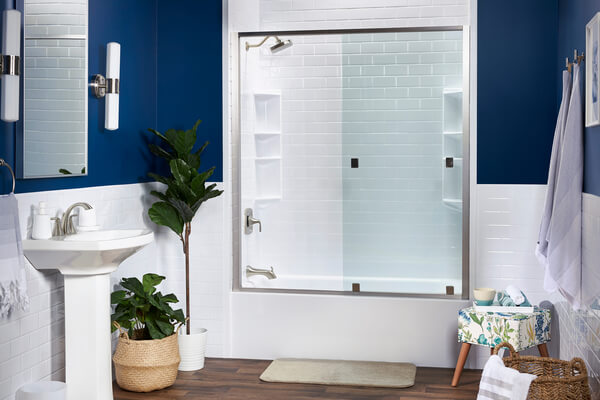 Bathroom with blue walls and white wainscoting featuring a white bathtub with subway tile walls and a glass door