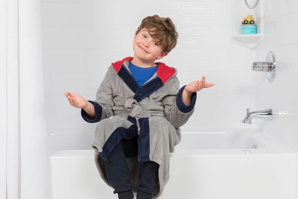 a boy wearing a bathrobe sitting on the ledge of a tub with his arms out