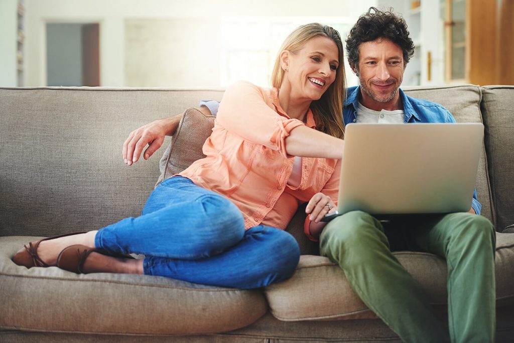 A woman sits on the floor with a man on the couch and his arms around her as they look at a laptop