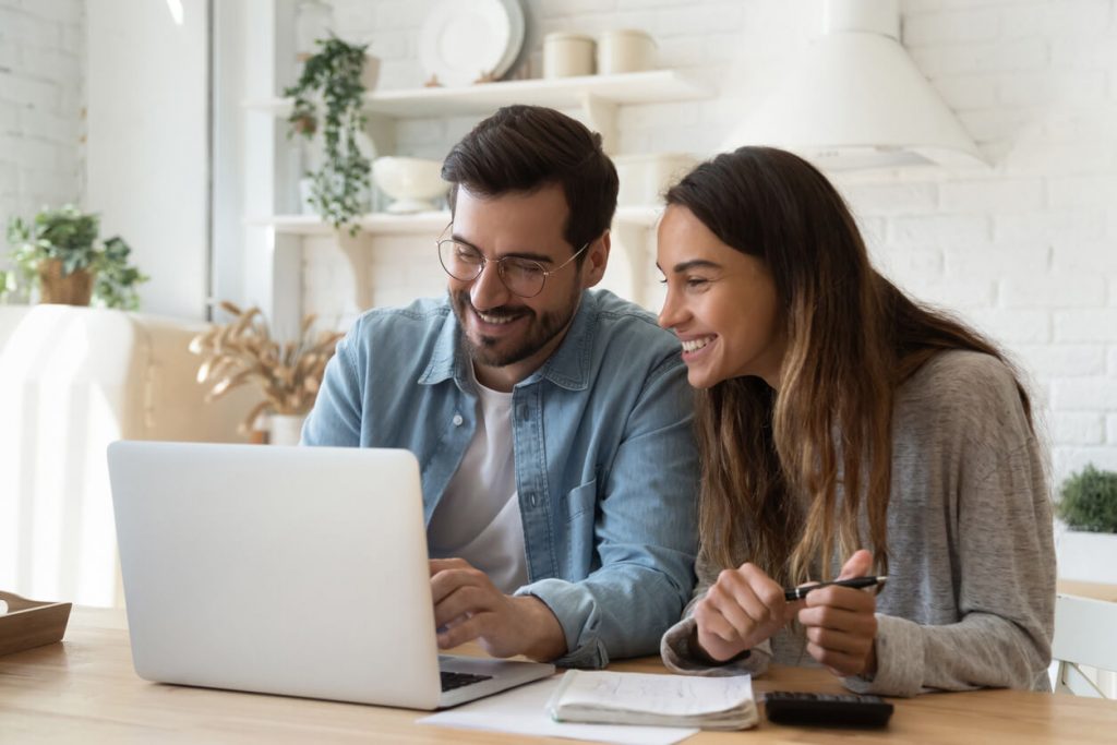 a man and woman sit at their kitchen table smiling over an opened laptop