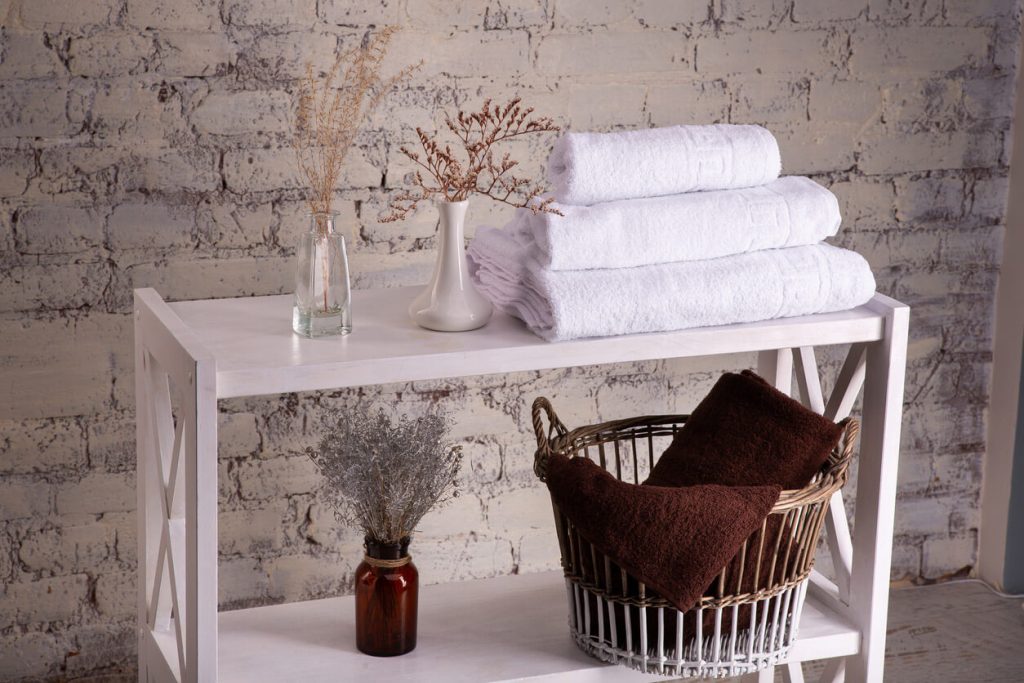 a white shelf stands against a brick wall holding white folded towels, basket containing a brown one, and vases with dried flowers