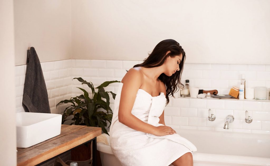 A woman wrapped in a towel sits on the edge of a tub in a relaxing styled bathroom feeling the water with her hand