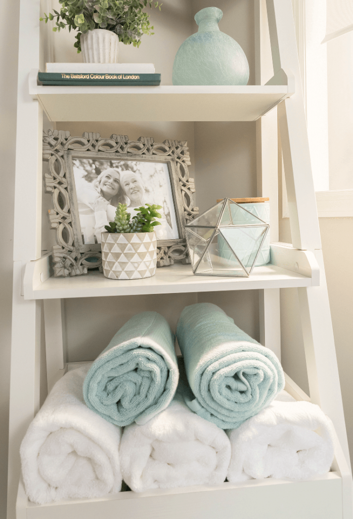 an open white ladder shelf with rolled up towels and framed pictures