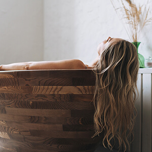A woman lays with her back towards the camera in a wooden tub in a modern bathroom