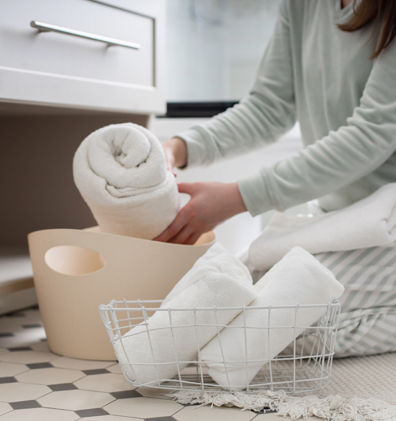 A woman is rolling towels into a basket under the vanity sink.