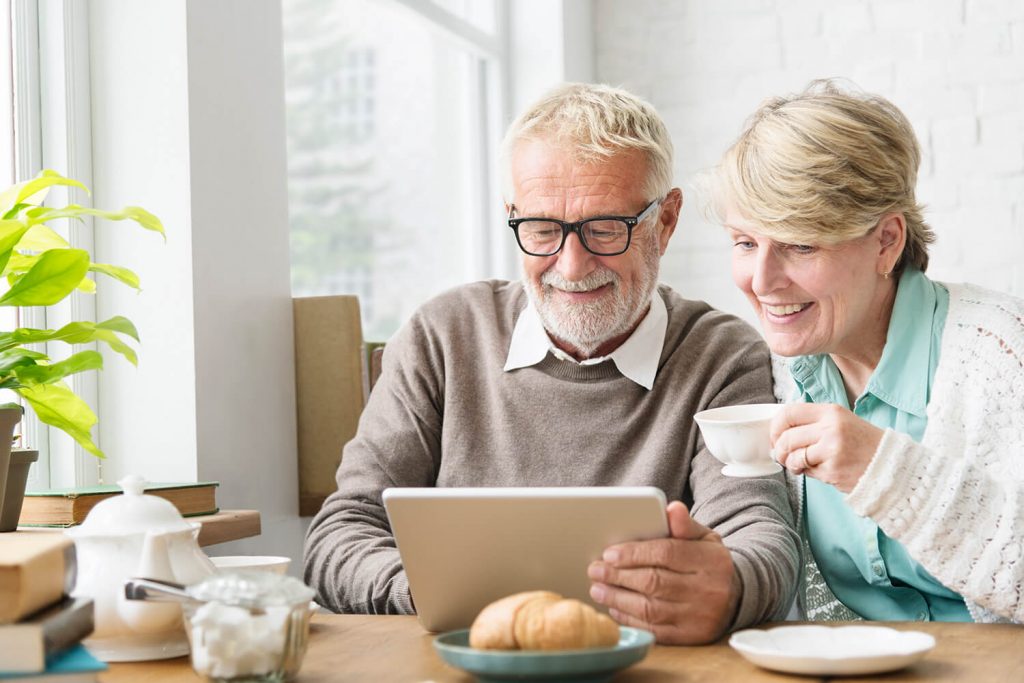 A mature couple sit together at a kitchen table smiling while looking at a tablet